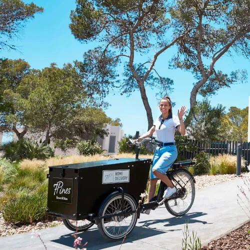 Frau fährt Fahrrad mit Anhänger, welcher den HOME DELIVERY Service von 7Pines Resort Ibiza anzeigt