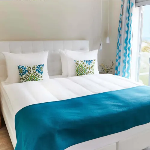 Spacious bedroom with a king-size bed, blue blanket, decorative pillows, and window with patterned curtains at 7Pines Resort Ibiza.