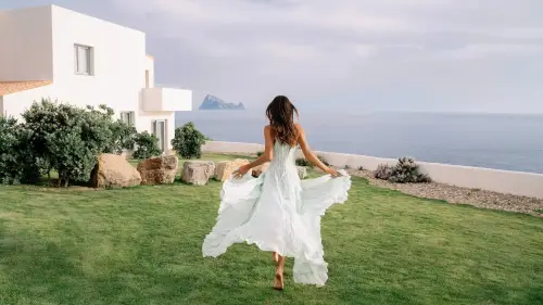 Woman in a flowing dress walking on a lawn near a white house with sea and sky in the background at 7Pines Resort Ibiza.